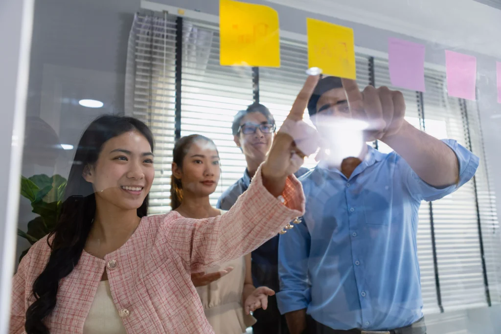 Profissionais apontando para post-its em um mural de vidro durante dinâmica de brainstorming, representando cultura de inovação.