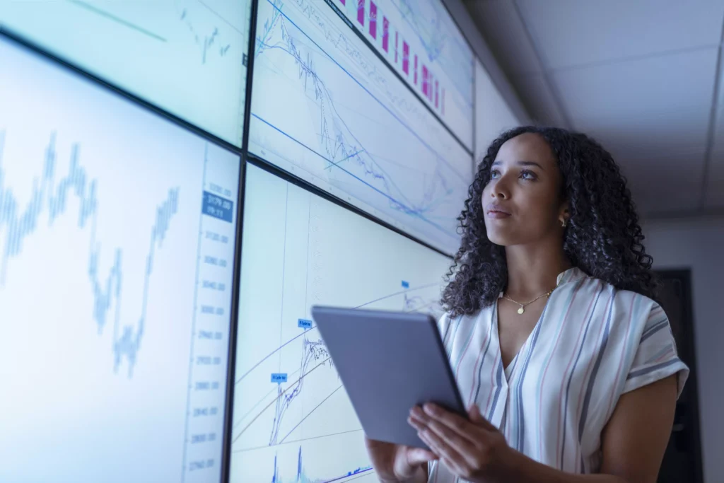 Mulher com tablet analisando painéis de dados exibidos em grandes telas, reforçando a estratégia data driven.