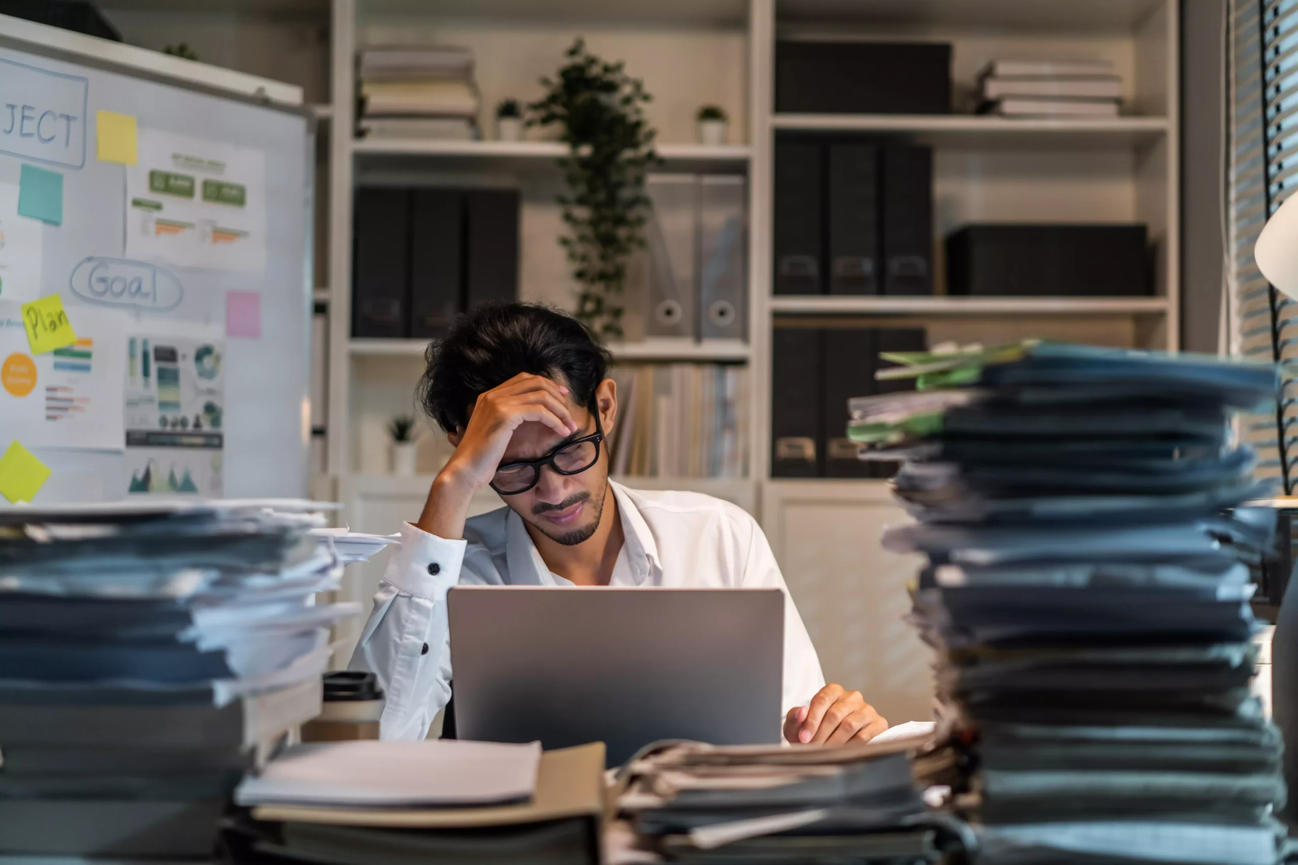 Homem estressado em frente ao computador, cercado por pilhas de documentos.
