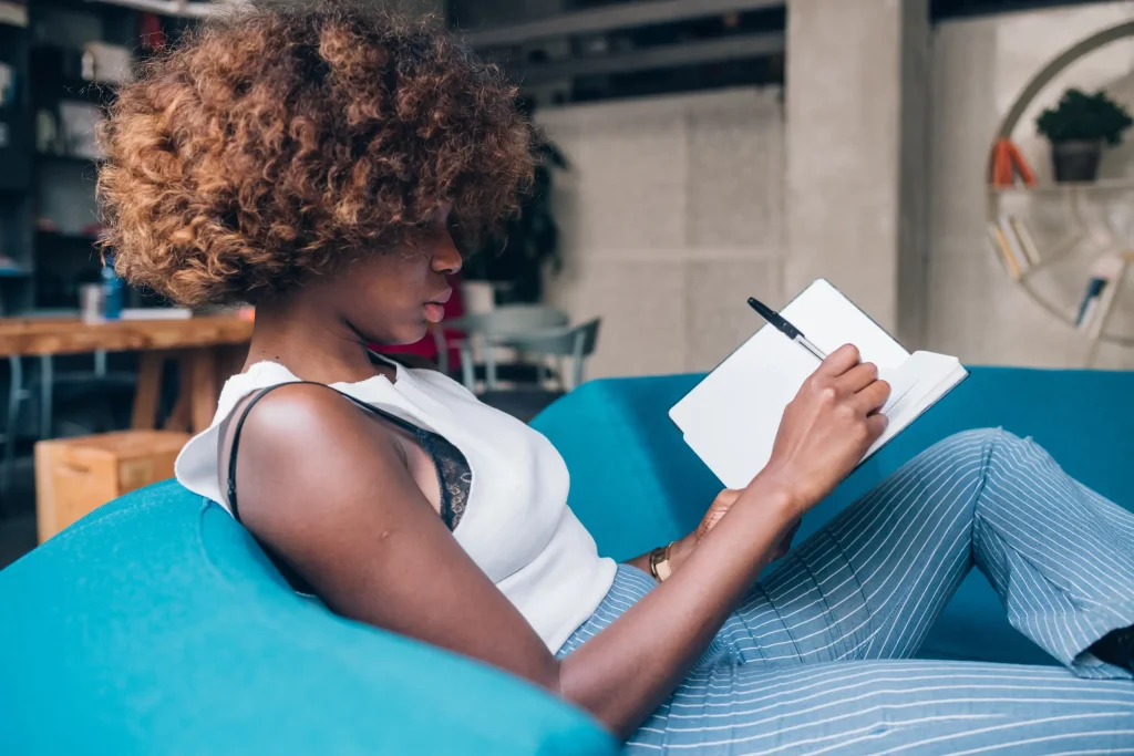 mulher negra sentada em um sofá azul escrevendo em um caderno|autoconhecimento|mulher asiática sorrindo em meio aos livros de uma prateleira|mulher negra sentada mexendo no computador