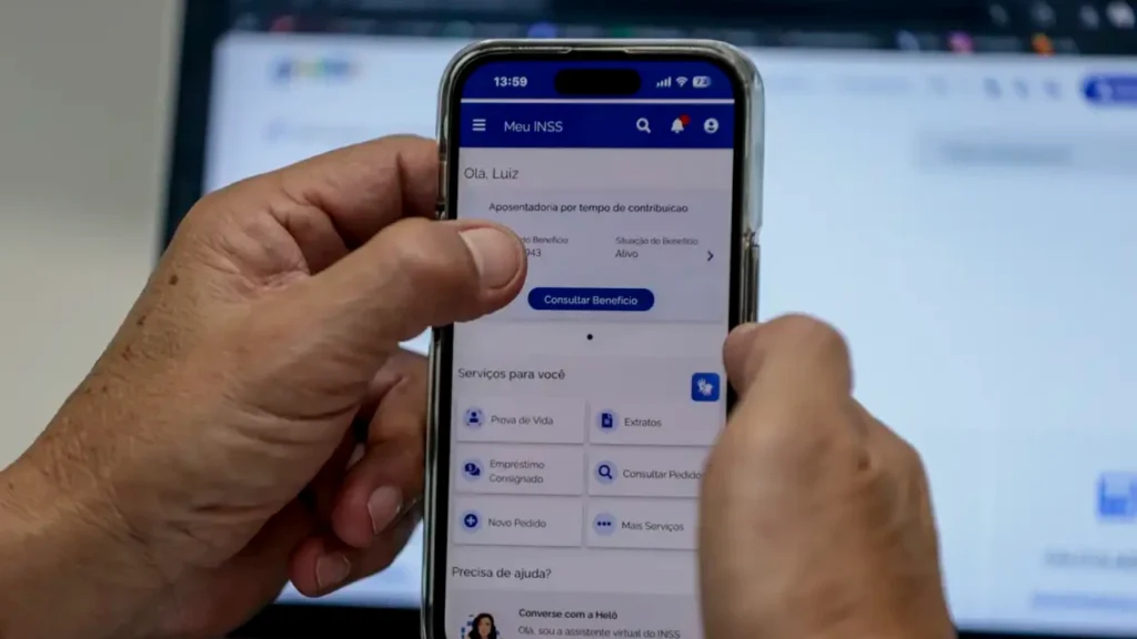 Uma pessoa segura um smartphone exibindo a tela inicial do aplicativo "Meu INSS", com opções como "Consultar Benefício" e "Serviços para você". Um computador com tela parcialmente visível está ao fundo.